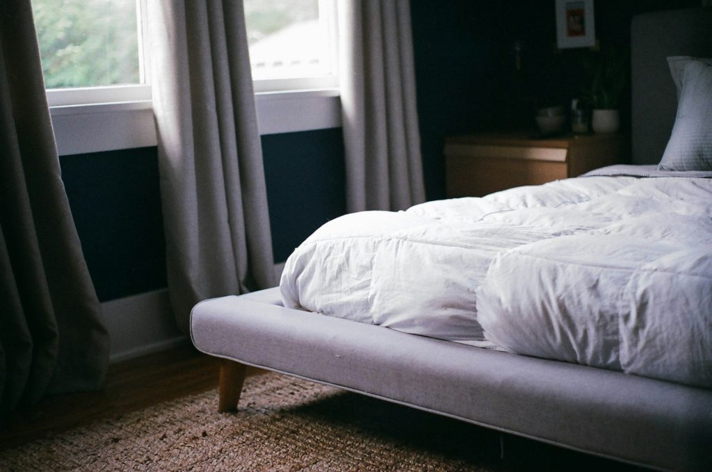 Bed met wit dekbed op een grijs gestoffeerd bedframe naast een raam
